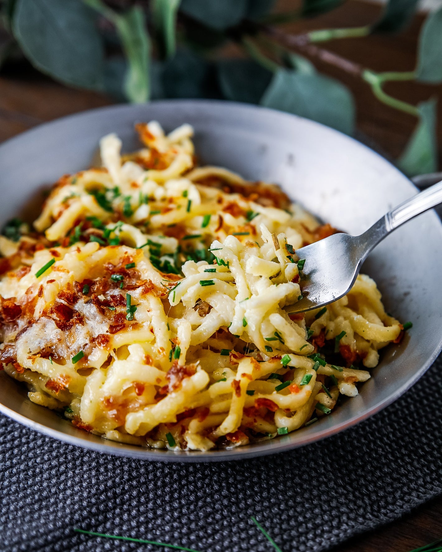 Eine Gabel hebt einen Bissen eines mit Schnittlauch garnierten Käse-Nudelgerichts in einer Schüssel auf einem strukturierten Tischset an.