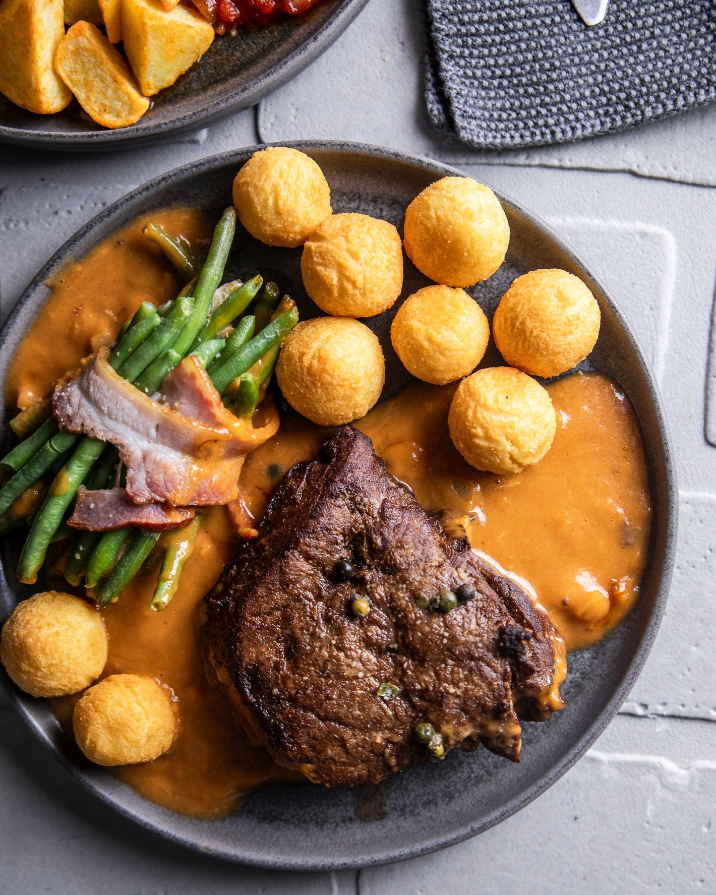 Ein Teller mit Steak, grünen Bohnen, Speck, runden Kartoffeln und brauner Soße auf einem grauen Tisch.