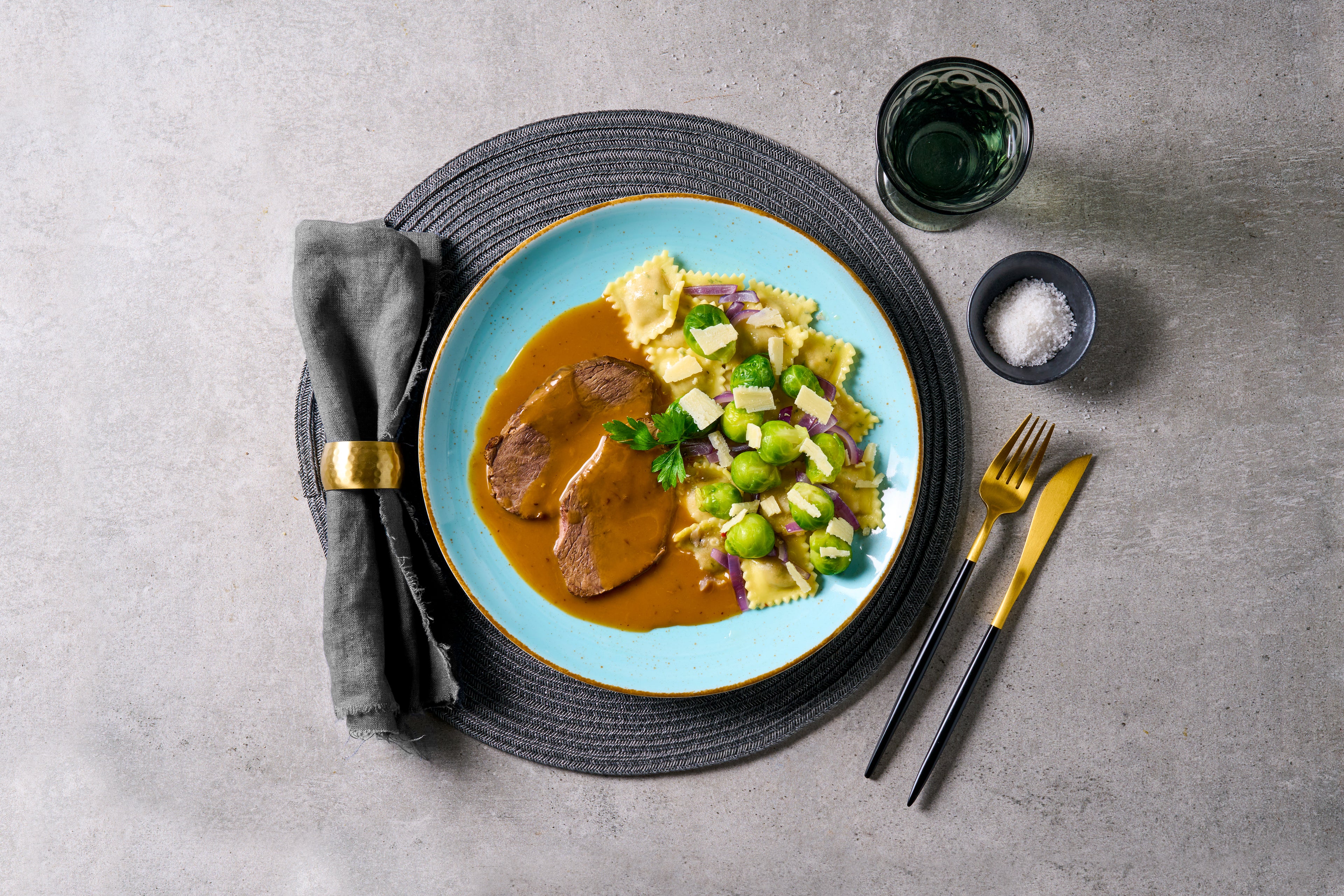 HOFMANNs Rinderbraten mit Hirschravioli auf einem blauen Teller mit Soße und Gemüse auf einer Tischunterlage.