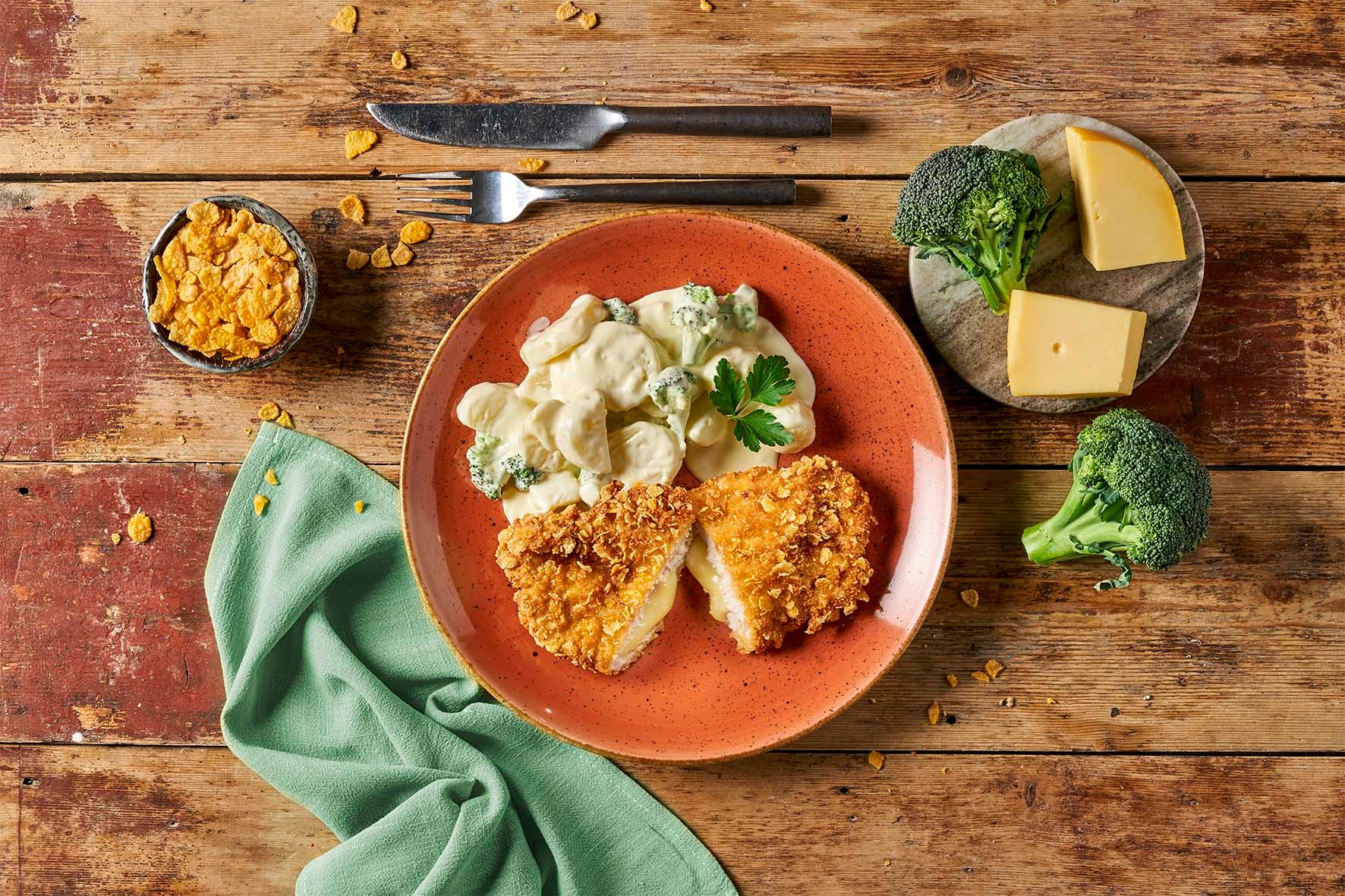 Ein Teller mit Käse-Kartoffelauflauf, Brokkoli und HOFMANNs Schnitzel-Box auf einem rustikalen Tisch.