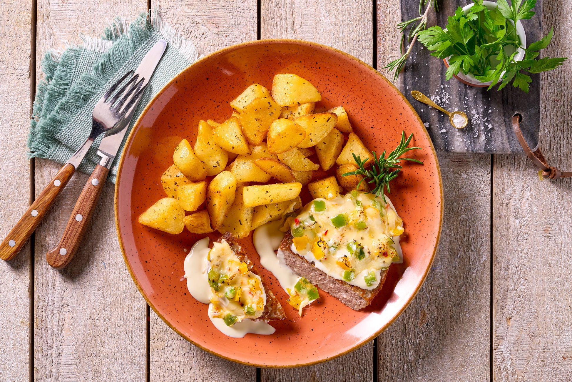Ein Teller mit Hofmanns Shop Hacksteak "Chili + Cheese", Patatas Bravas und Rosmarin auf einem hellen Tisch.