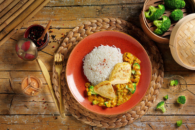 Ein roter Teller mit weißem Reis, Tofu und Gemüse auf einer gewebten Matte, neben Schüsseln mit Paprika und Brokkoli.