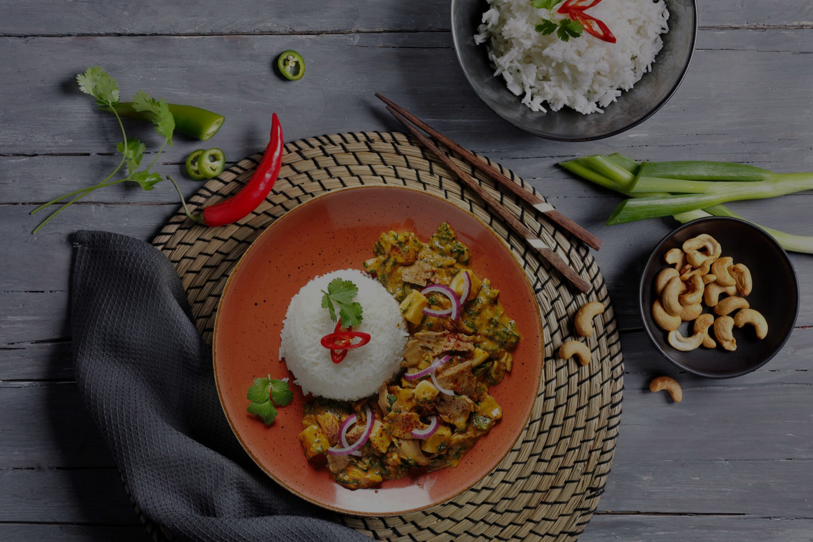 Ein Teller mit Reis, Curry und Garnitur, dazu eine Schüssel Reis, Cashews und Essstäbchen auf einer gewebten Matte.
