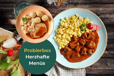 Zwei Teller von HOFMANNs Herzhafte Küche bieten kulinarischen Hochgenuss auf einem rustikalen Holztisch.