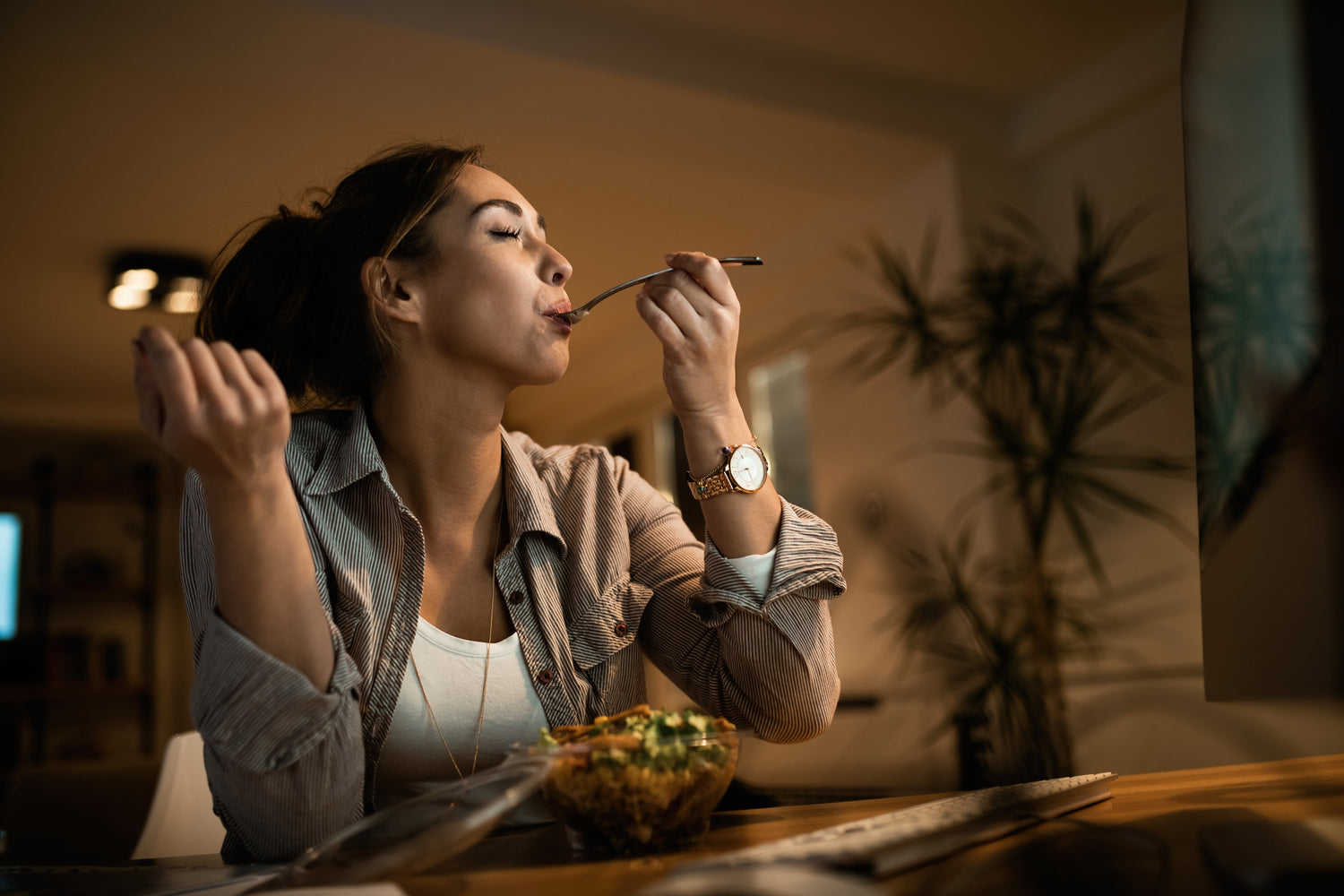 Frau genießt eine Mahlzeit zu Hause, hält mit geschlossenen Augen eine Gabel, eine Salatschüssel und eine aufgeschlagene Zeitschrift vor sich.