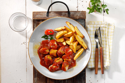 Ein Teller HOFMANNs Neuheiten Currywurst mit Pommes, garniert mit Petersilie, serviert mit Besteck.