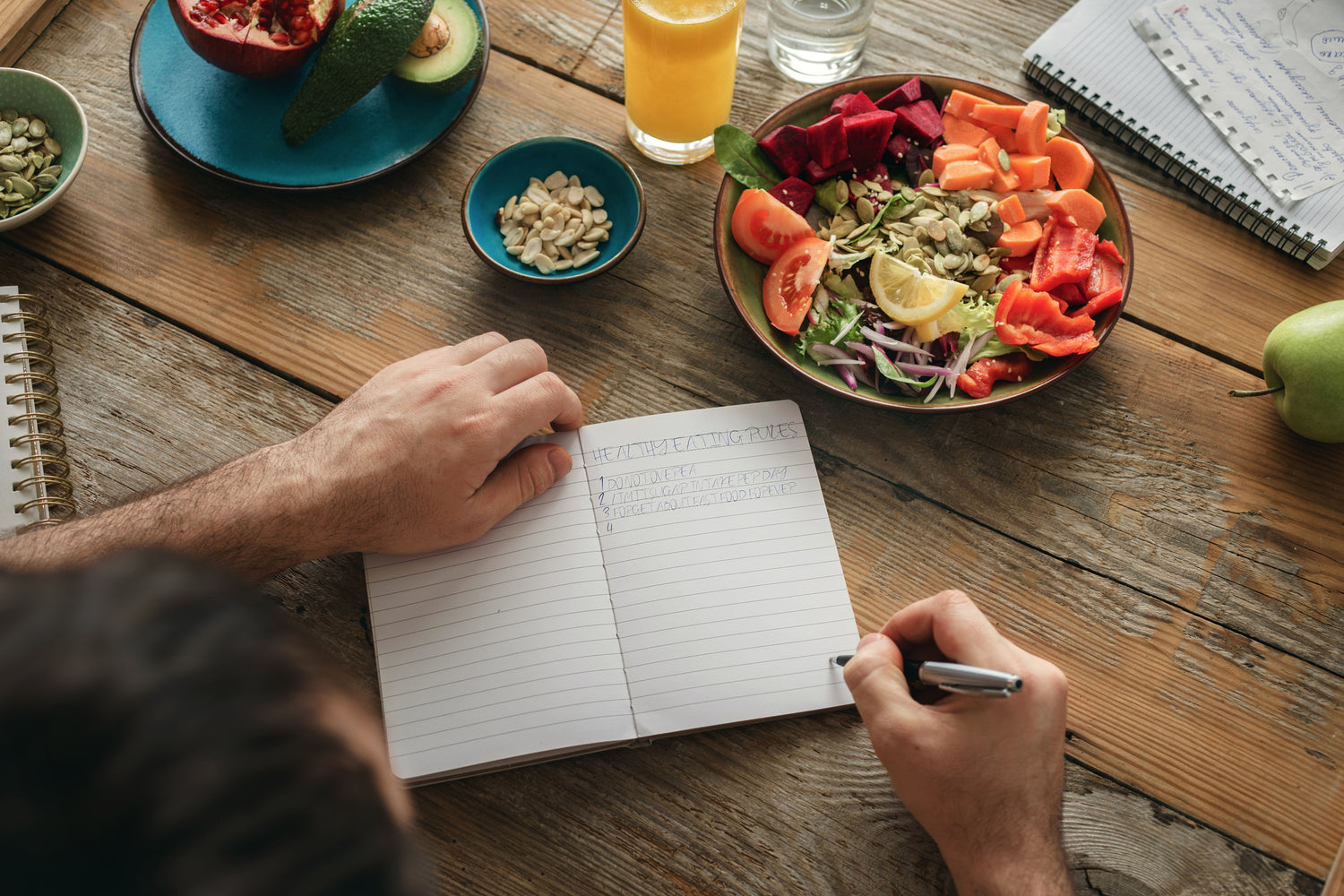Person, die in ein Notizbuch schreibt, auf dem ein bunter Salat, Samen, Obst und ein Glas Saft auf einem Holztisch stehen.