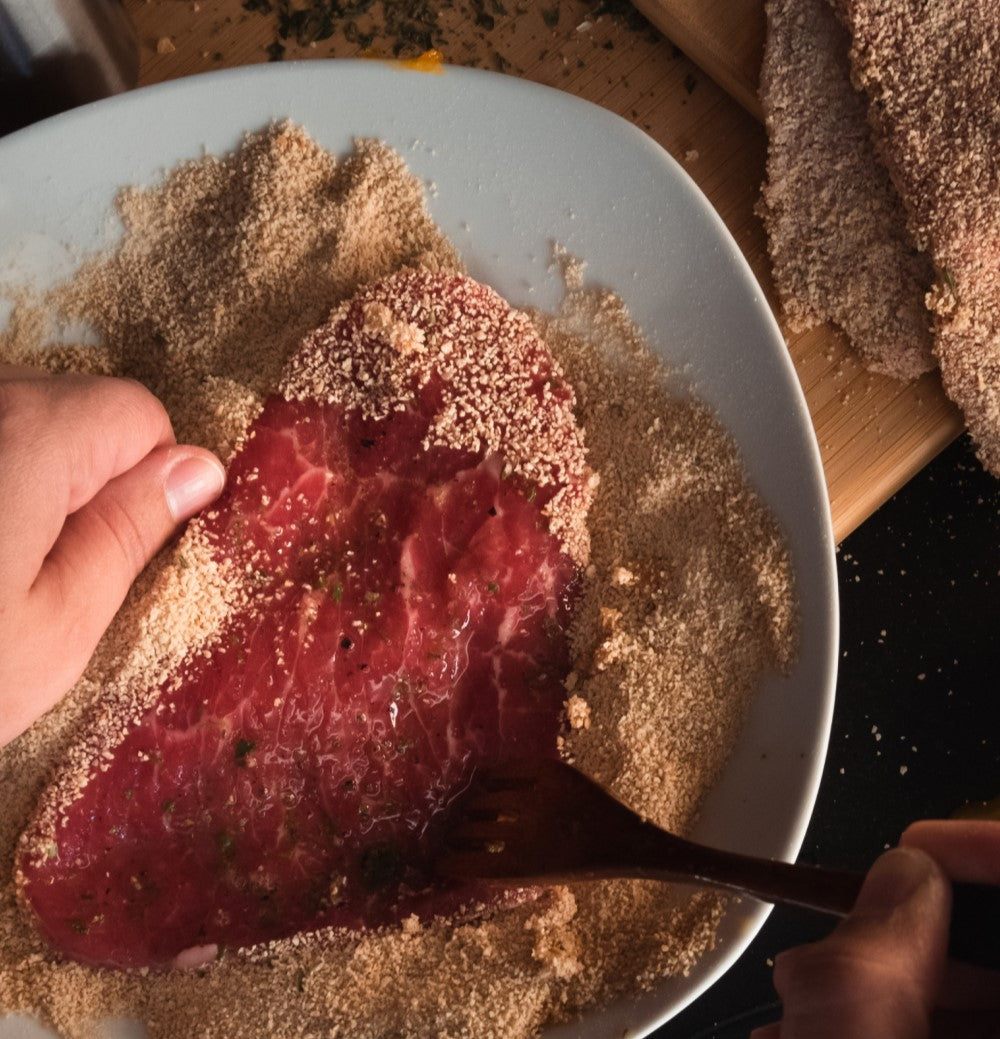 Nahaufnahme von Händen, die ein rohes Schnitzel auf einem weißen Teller mit Semmelbröseln panieren.