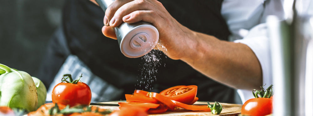 Person würzt geschnittene Tomaten auf einem Schneidebrett, umgeben von anderem frischen Gemüse.
