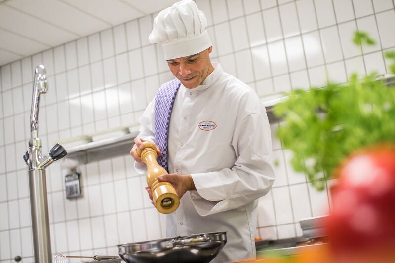 Ein Koch würzt in einer Küche das Essen. Er trägt eine weiße Kochjacke und -mütze, im Vordergrund sieht man buntes Gemüse.