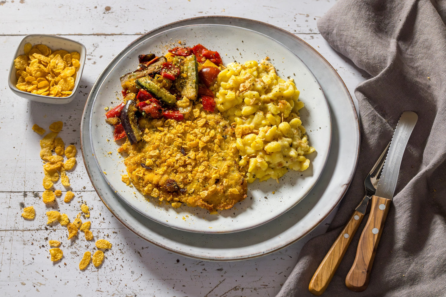Teller mit paniertem gebratenem Fleisch, Makkaroni und Käse, geröstetem Gemüse, Besteck und verschütteten Cornflakes als Beilage.