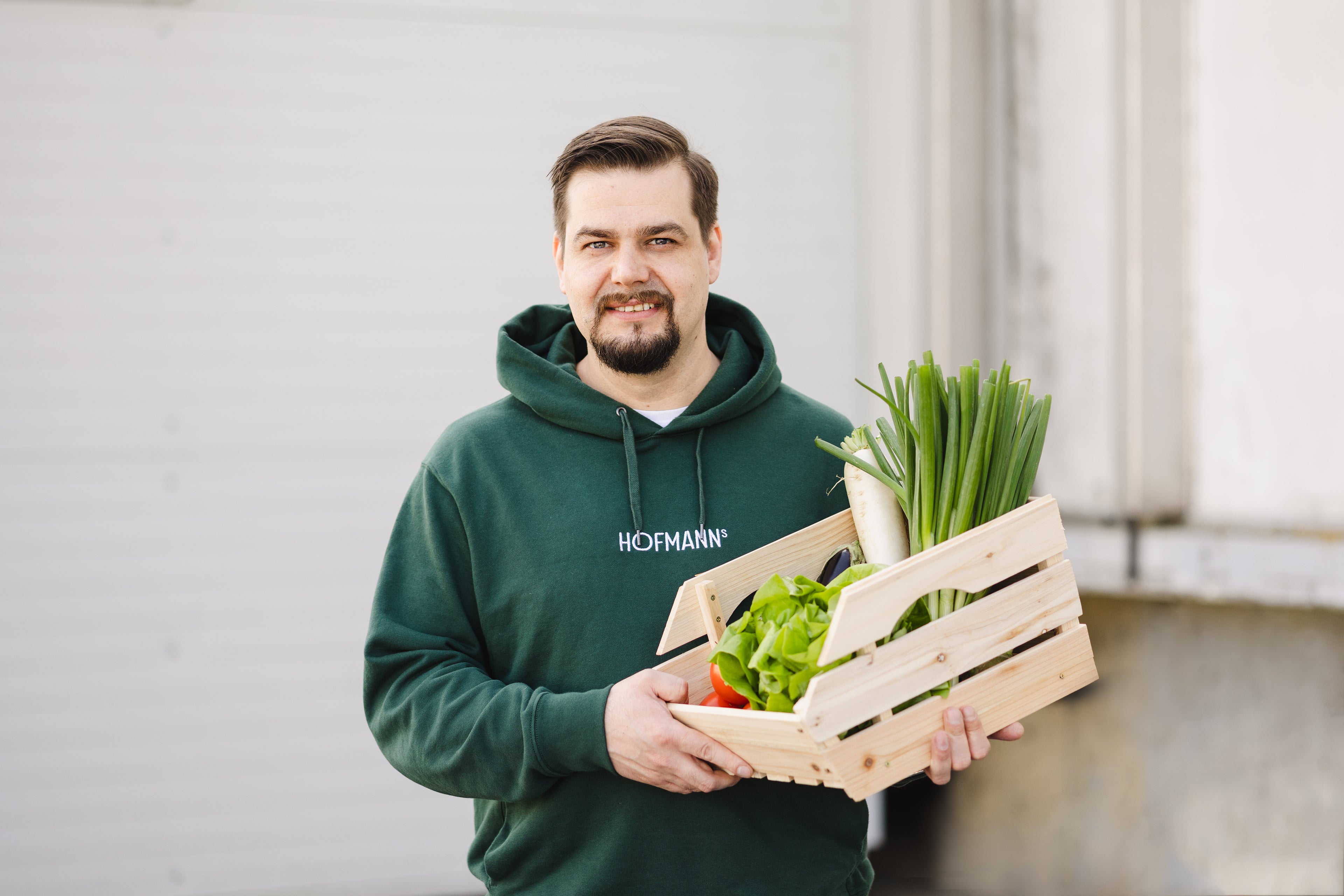 Mann im grünen Sweatshirt hält Holzkiste mit frischem grünem Gemüse und lächelt vor hellem Hintergrund.