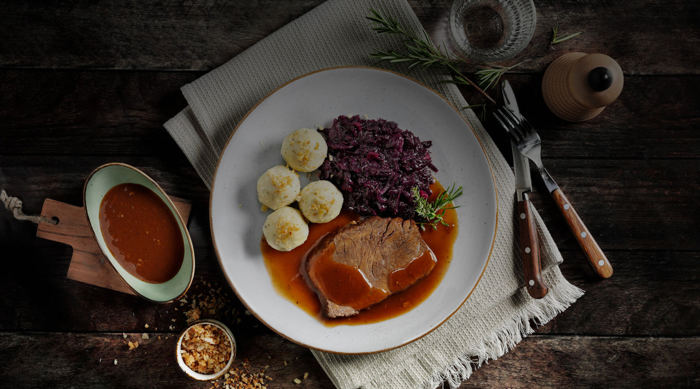 Ein Teller Roastbeef mit Bratensoße, gekochten Knödeln und Rotkohl, garniert mit Kräutern, dazu Soße.