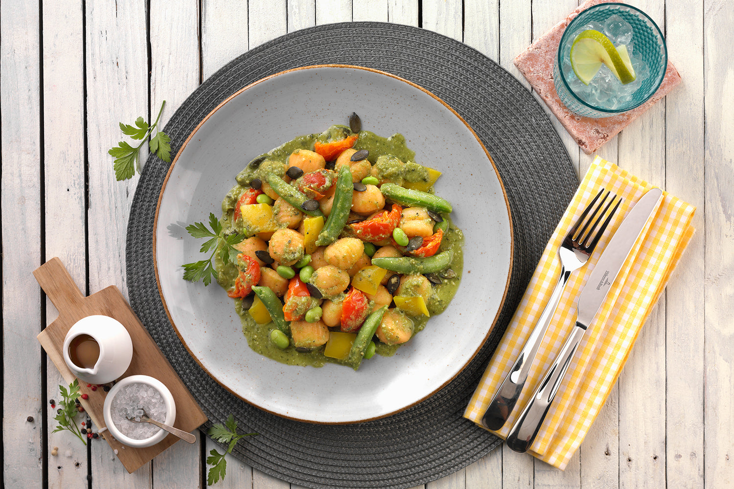 Ein Teller mit bunten Gnocchi mit Gemüse in grüner Soße, daneben Besteck und ein Glas mit Zitronenscheibe.