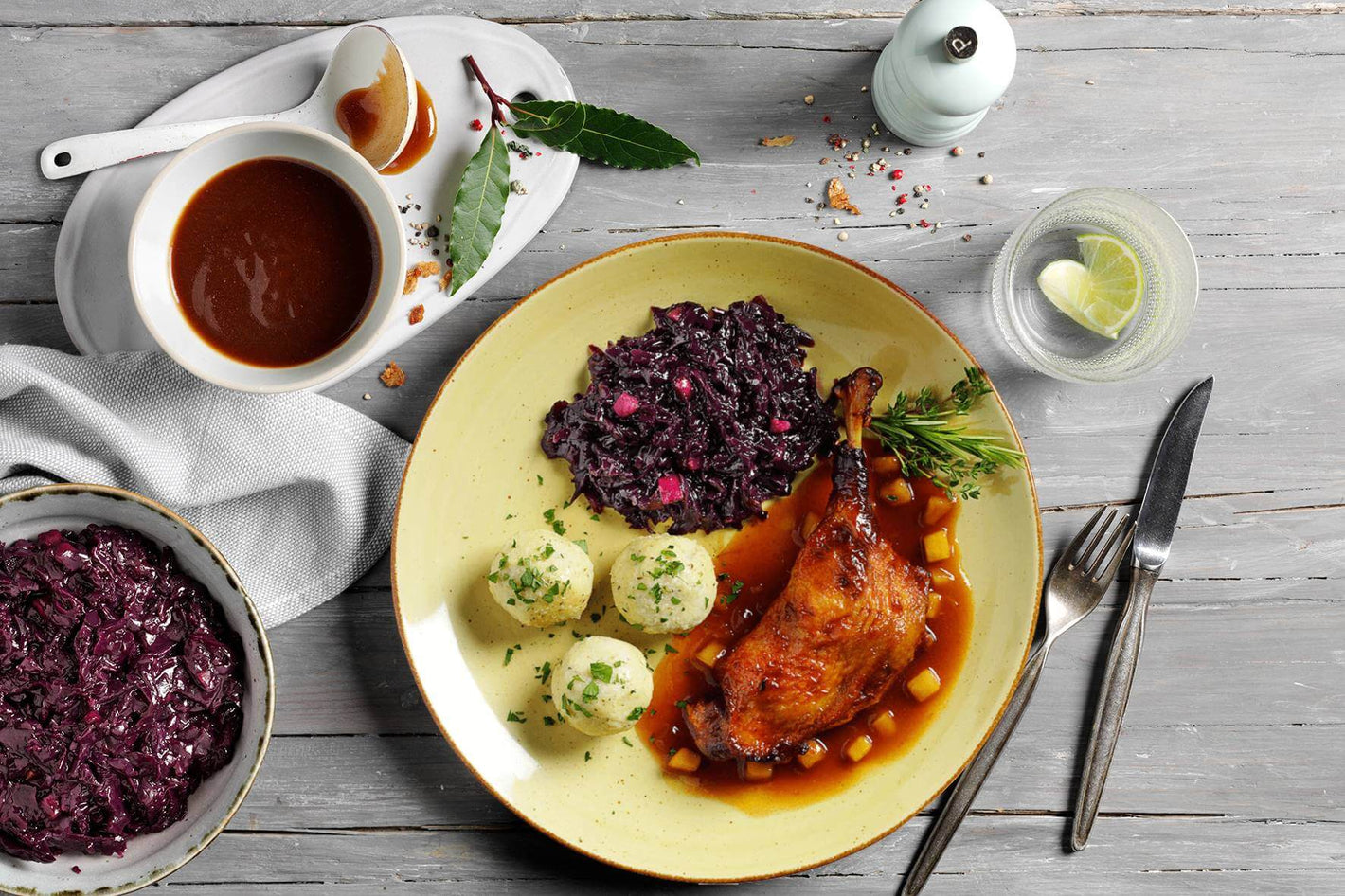 Teller mit gebratener HOFMANNs Entenkeule, Bratapfelsoße, Kartoffelknödel und Apfelrotkohl auf einem Holztisch, Getränk mit Zitrone von Hofmanns Shop.