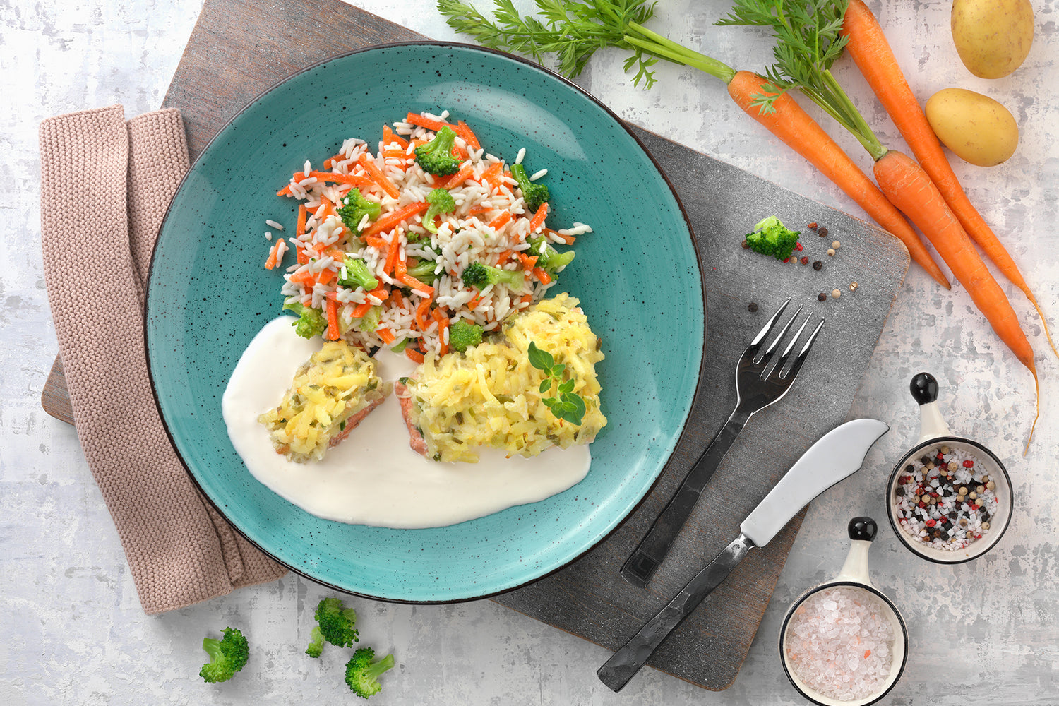 Auf einem blaugrünen Teller liegt Lachs mit Kartoffelkruste von Hofmanns Shop, garniert mit einer cremigen Weißweinsoße und einer Prise Kräutern. Besteck liegt in der Nähe.