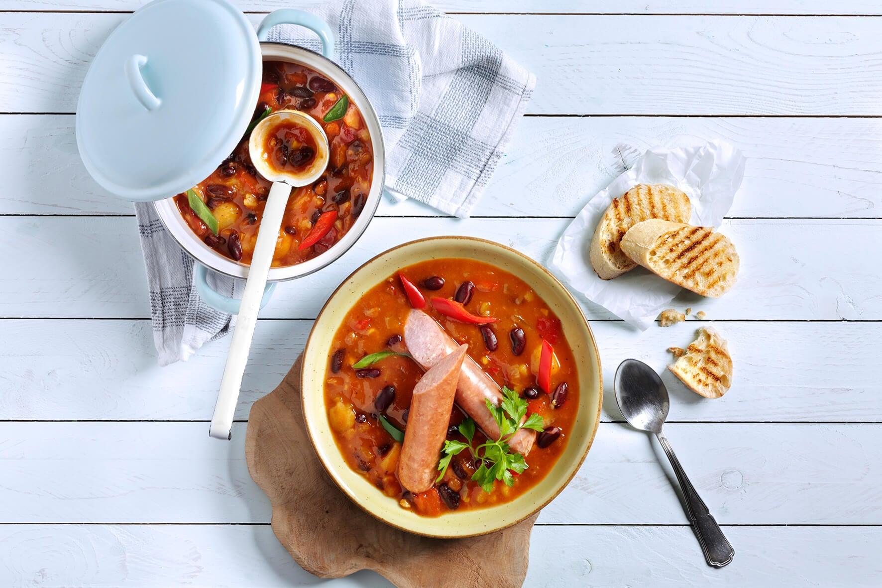 Schüssel mit HOFMANNs Suppen & Eintöpfe mit Bohnen, Wurst und Brot auf einem weißen Holztisch.