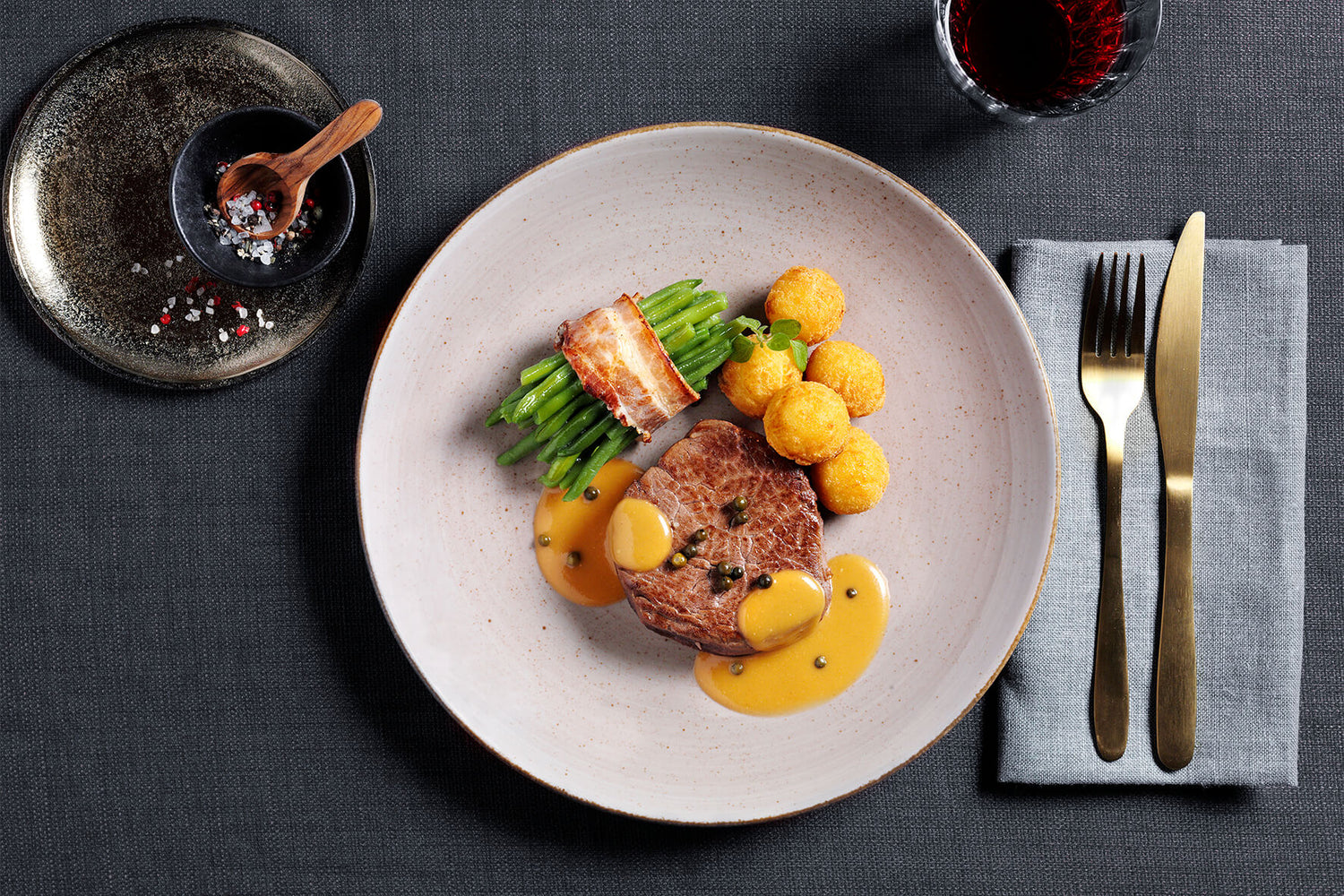 Gourmet-Menü mit Steak, Bohnen, Käsebällchen und Soße auf einem weißen Teller, neben Besteck und Rotwein.