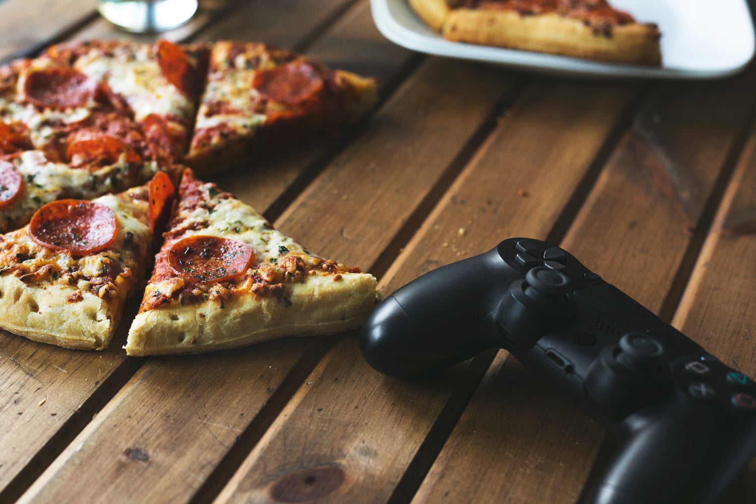 Pizzastücke mit Peperoni auf einem Holztisch, daneben ein Gamecontroller.