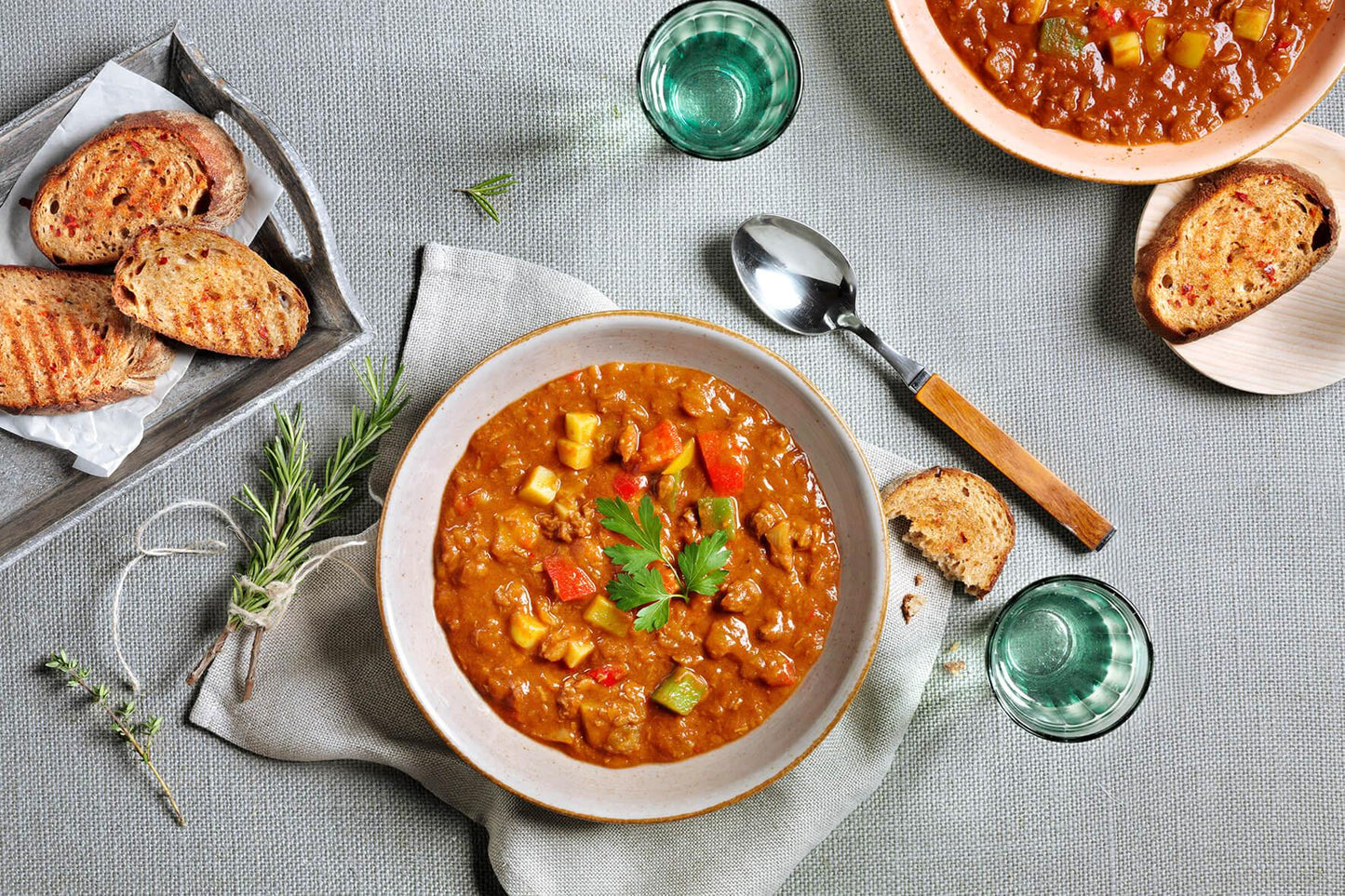 Gulaschsuppe „Ungarische Art“ von Hofmanns Shop serviert mit Brot und Wasser auf einer grauen Tischdecke.