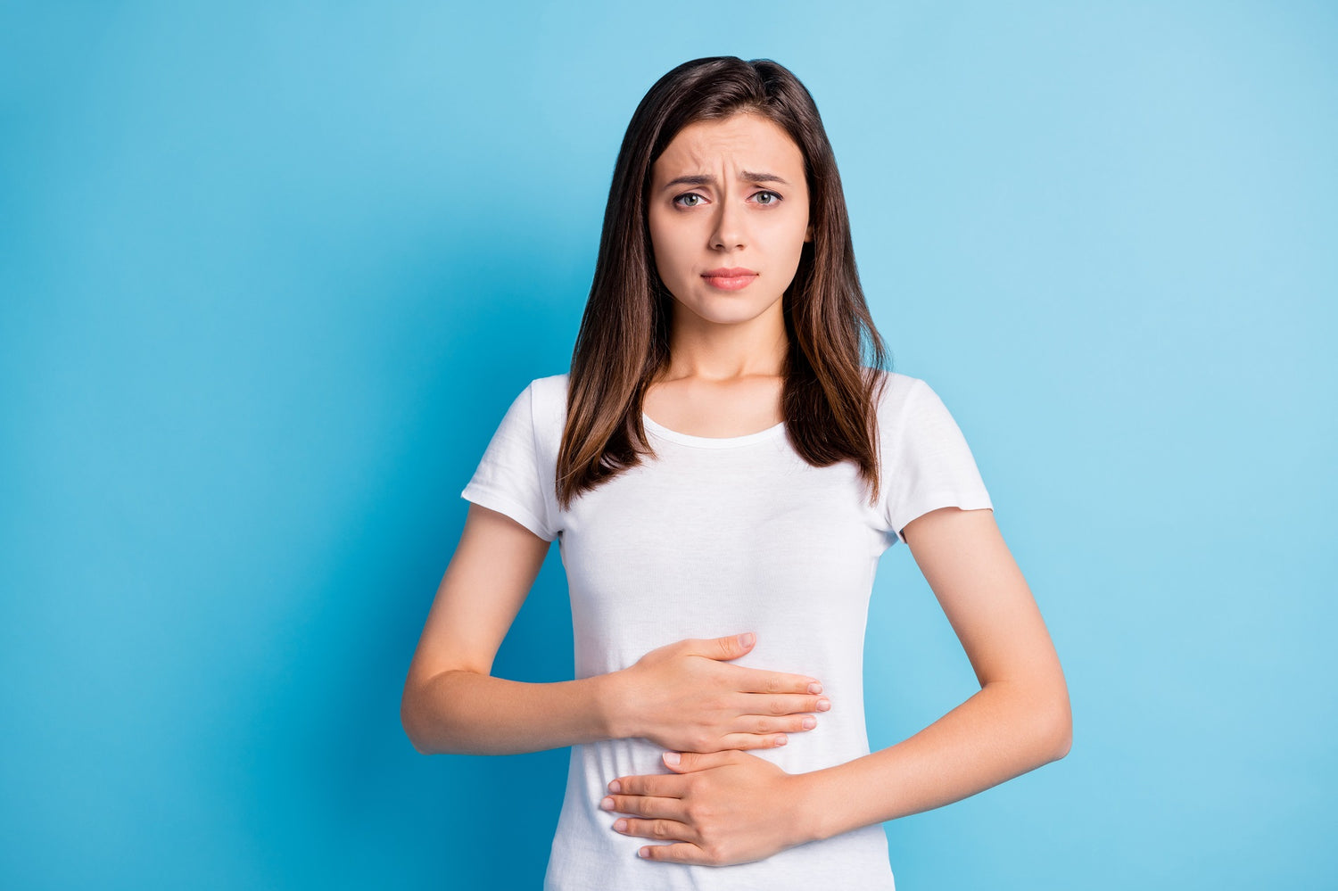 Frau mit braunen Haaren in weißem Hemd vor blauem Hintergrund, hält sich mit schmerzverzerrtem Gesichtsausdruck den Bauch.