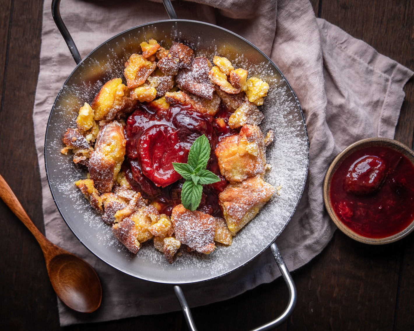 Eine Schüssel Kaiserschmarrn mit Puderzucker und Beerensauce, garniert mit Minze. Holzlöffel daneben.