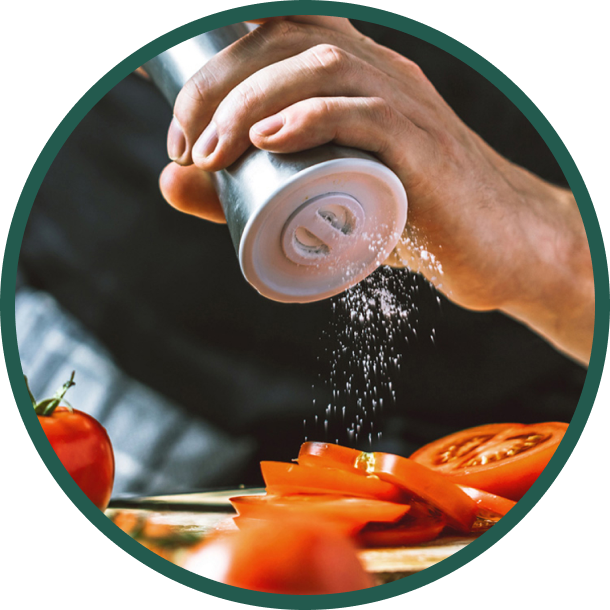 Hände streuen mit einem Salzstreuer Salz auf Tomatenscheiben, im Hintergrund sind ganze Tomaten zu sehen.