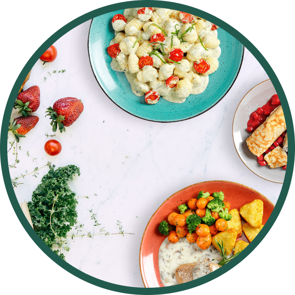 Teller mit Gnocchi, Gemüse und Erdbeerdessert sind auf einem Tisch angerichtet, überall herum liegen frische Produkte.