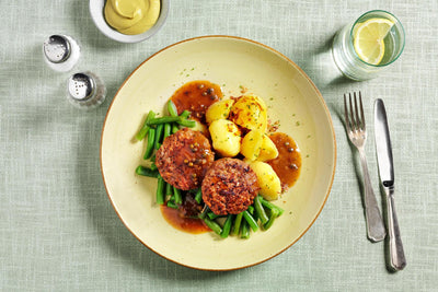 Eine HOFMANNs Feinschmecker-Box: zwei Patties, Kartoffeln, grüne Bohnen & braune Sauce auf einem grünen Tuch.