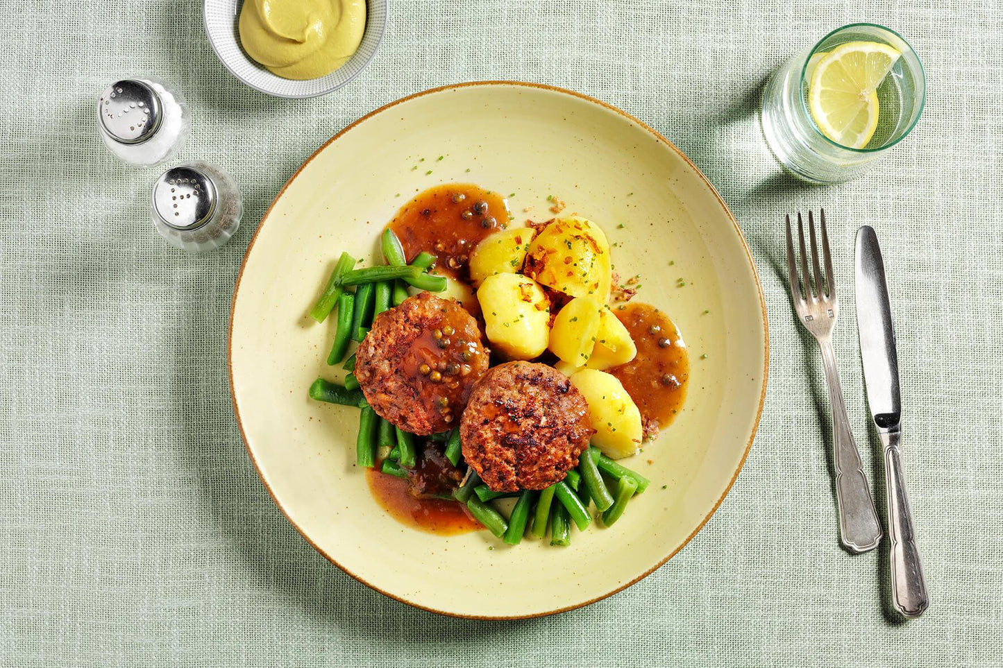 Eine HOFMANNs Feinschmecker-Box: zwei Patties, Kartoffeln, grüne Bohnen & braune Sauce auf einem grünen Tuch.