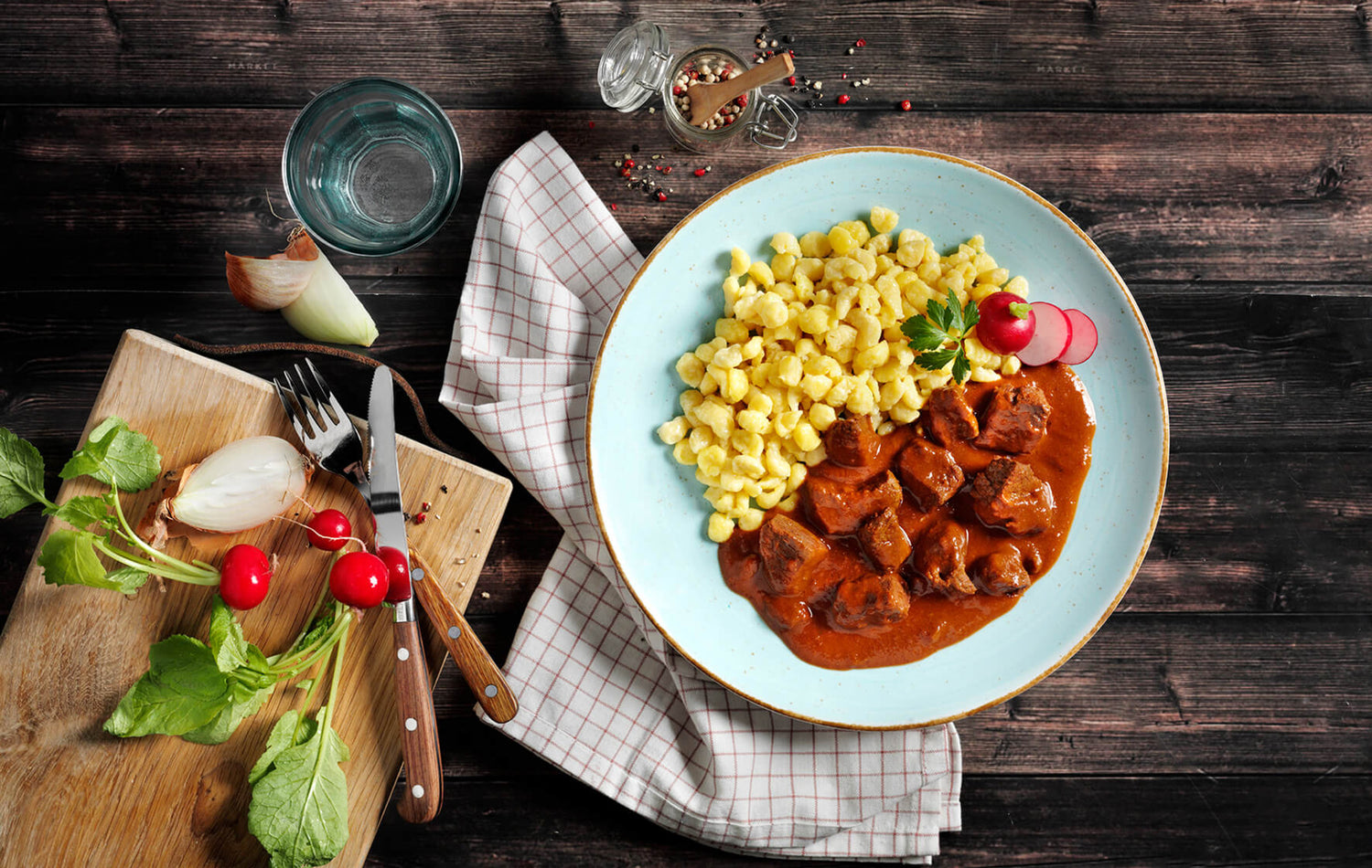 Teller mit Eintopf und Nudeln auf einem Tisch mit Salat, Besteck und Gewürzen auf einer rustikalen Holzoberfläche.