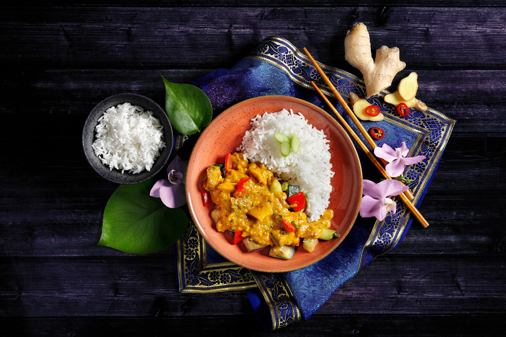 Ein Gericht aus Reis und Curry mit Gemüse, garniert mit Frühlingszwiebeln, perfekt für ein romantisches Abendessen auf einem dunklen Tisch, geschmückt mit Blättern und Ingwer aus der HOFMANNs Valentinstags-Box von Hofmanns Shop.