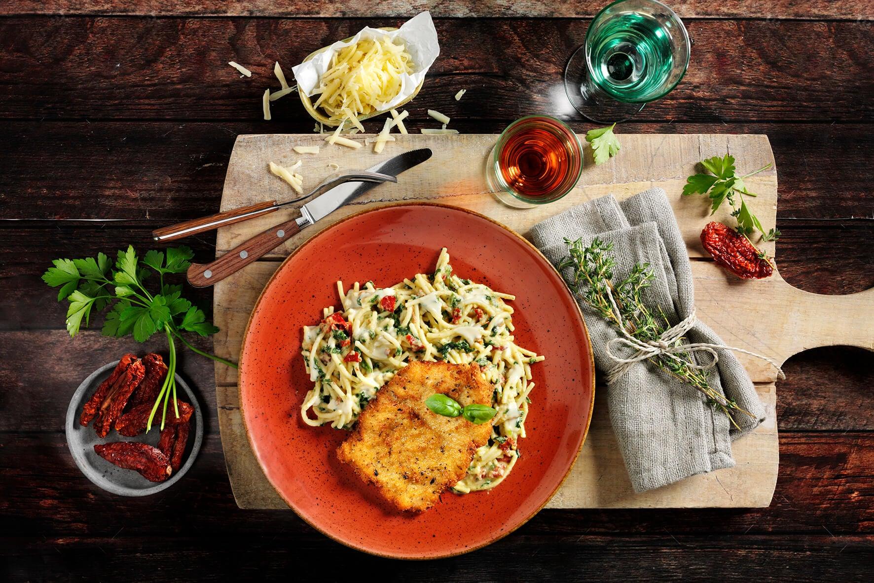 HOFMANNs Schnitzel-Box + „Schwäbische Spezialitäten“ mit Pasta, Käse & Wein auf rustikalem Tisch.