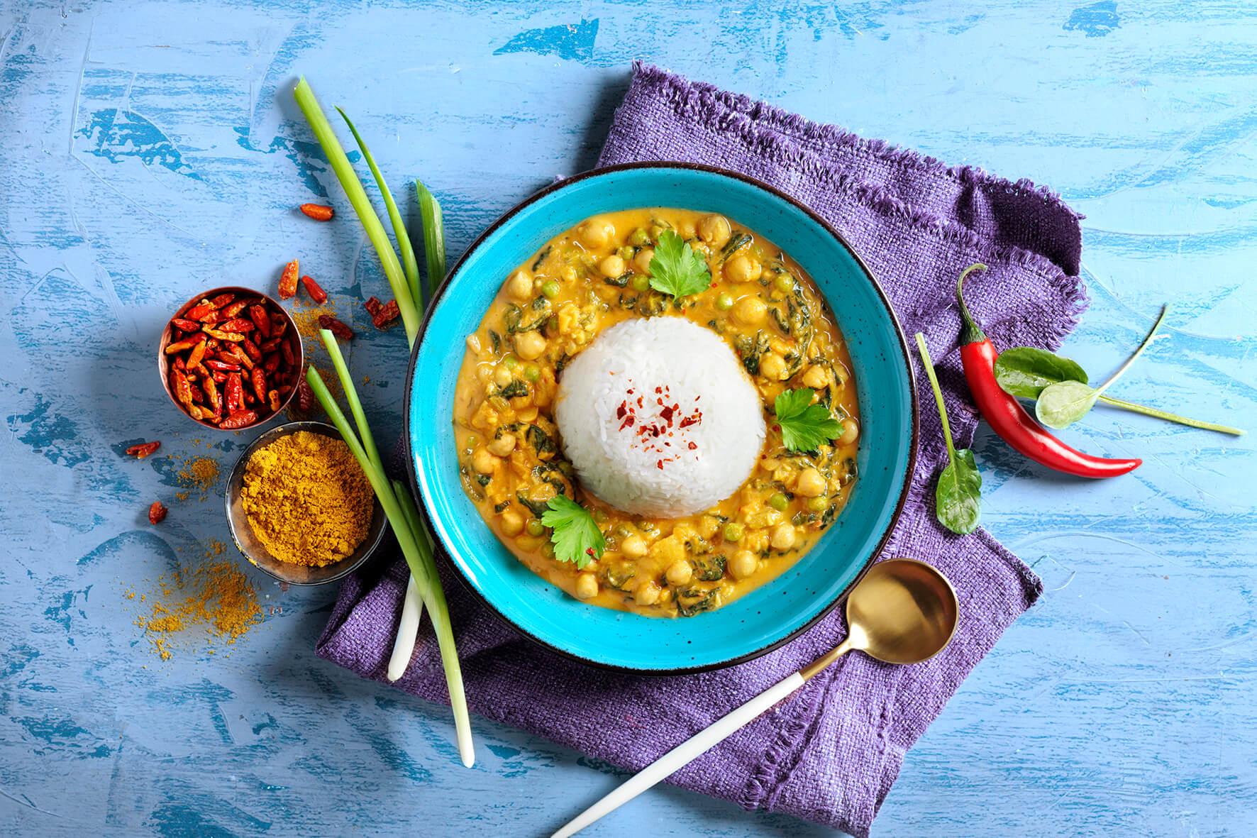 Eine Schüssel Curry mit Reis aus der HOFMANNs Vegan-Box neben Gewürzen und Frühlingszwiebeln auf einem blauen Tisch.