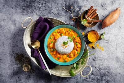 Süßkartoffel-Curry von Hofmanns Shop mit Basmatireis, umgeben von Gewürzen und Süßkartoffel.