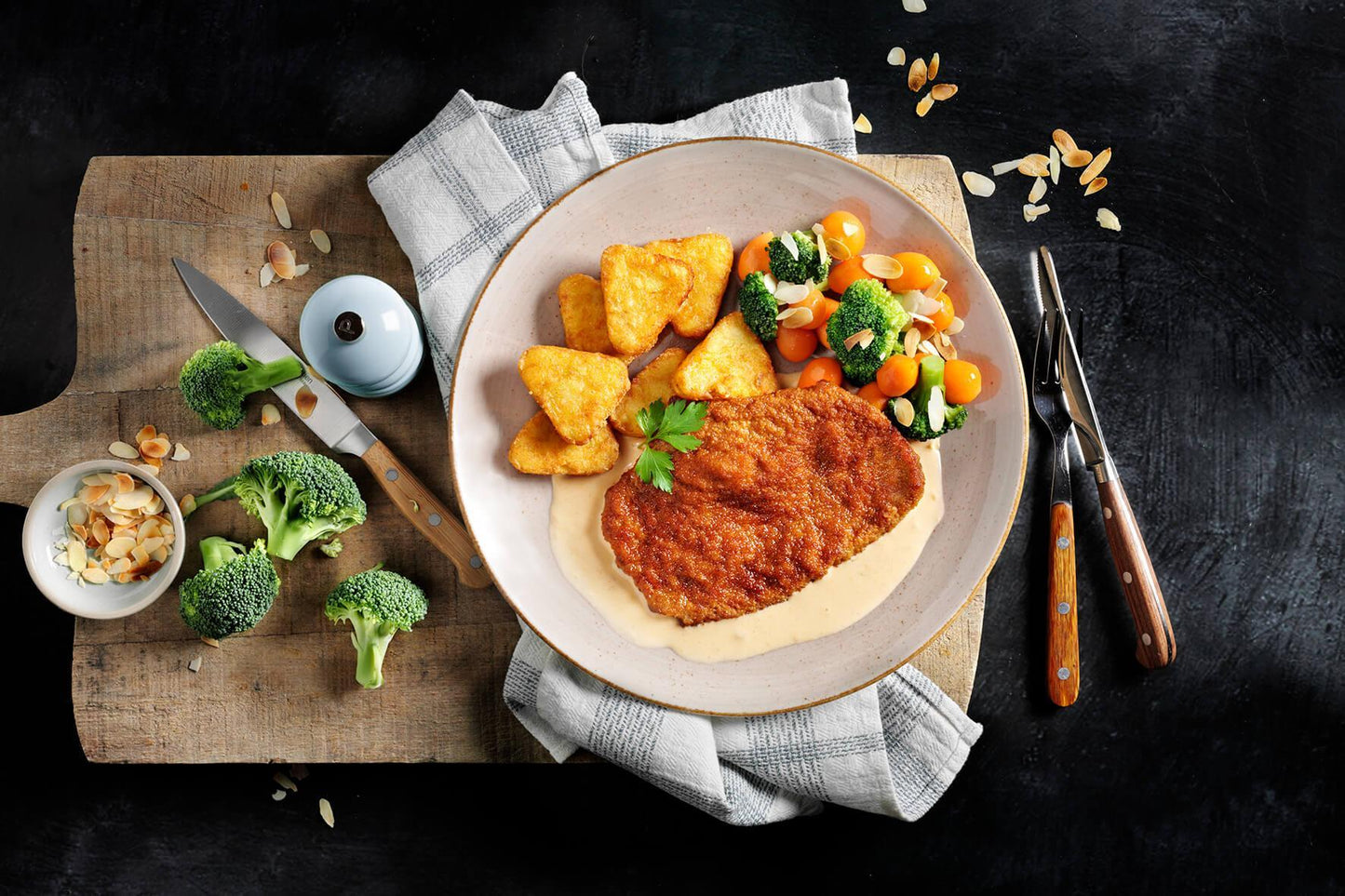 HOFMANNs Deutscher Klassiker: paniertes Schnitzel, Sauce, Wedges & Gemüse serviert auf einem Holzbrett.