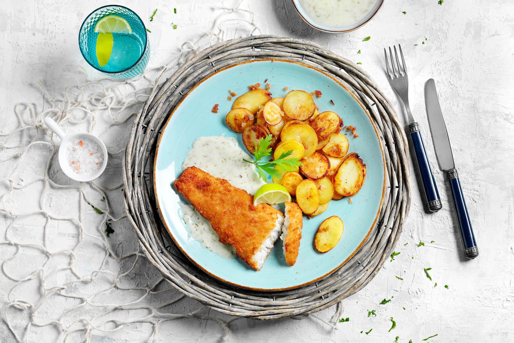 Ein Teller mit Hofmanns Shop Kabeljaufilet „Kräuter & Speck“, Bratkartoffeln, Dip, Zitrone und einem Getränk.