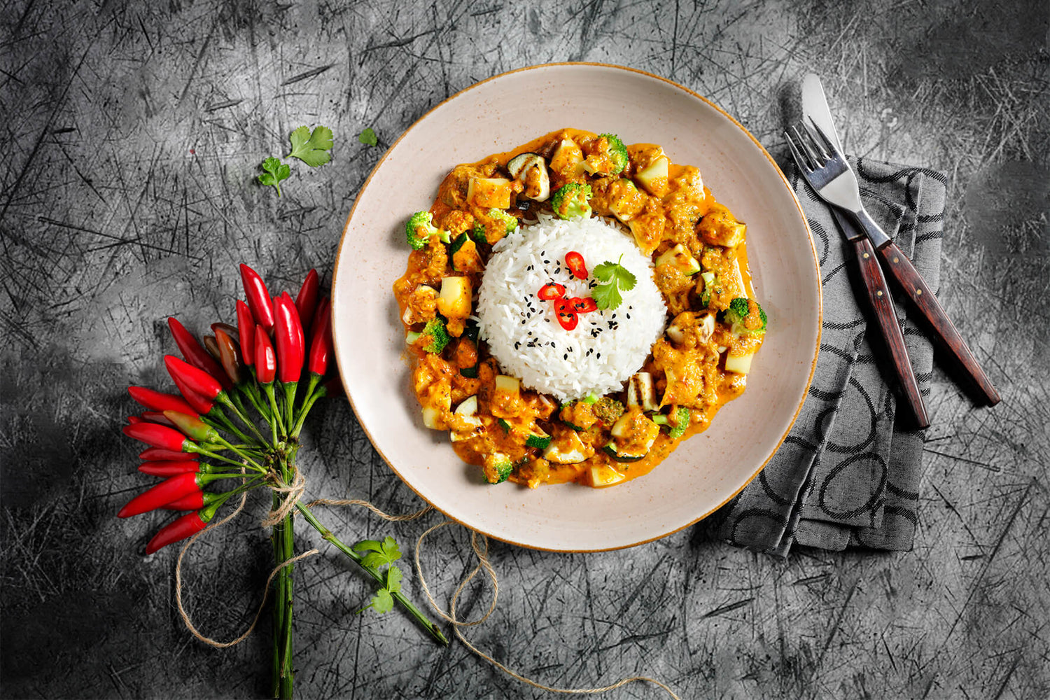 Ein Teller Reis und Curry mit Tofu und Gemüse, garniert mit roten Chilis, steht neben roten Chilischoten und Besteck – eine exquisite Hofmanns Vegan-Box, die das Beste der asiatischen Küche präsentiert.