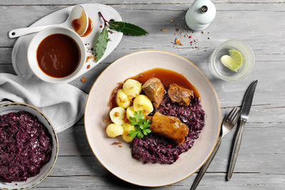HOFMANNs Hausmannskost-Box: Rinderrouladen, Knödel, Rotkohl & Soße auf einem rustikalen Tisch.
