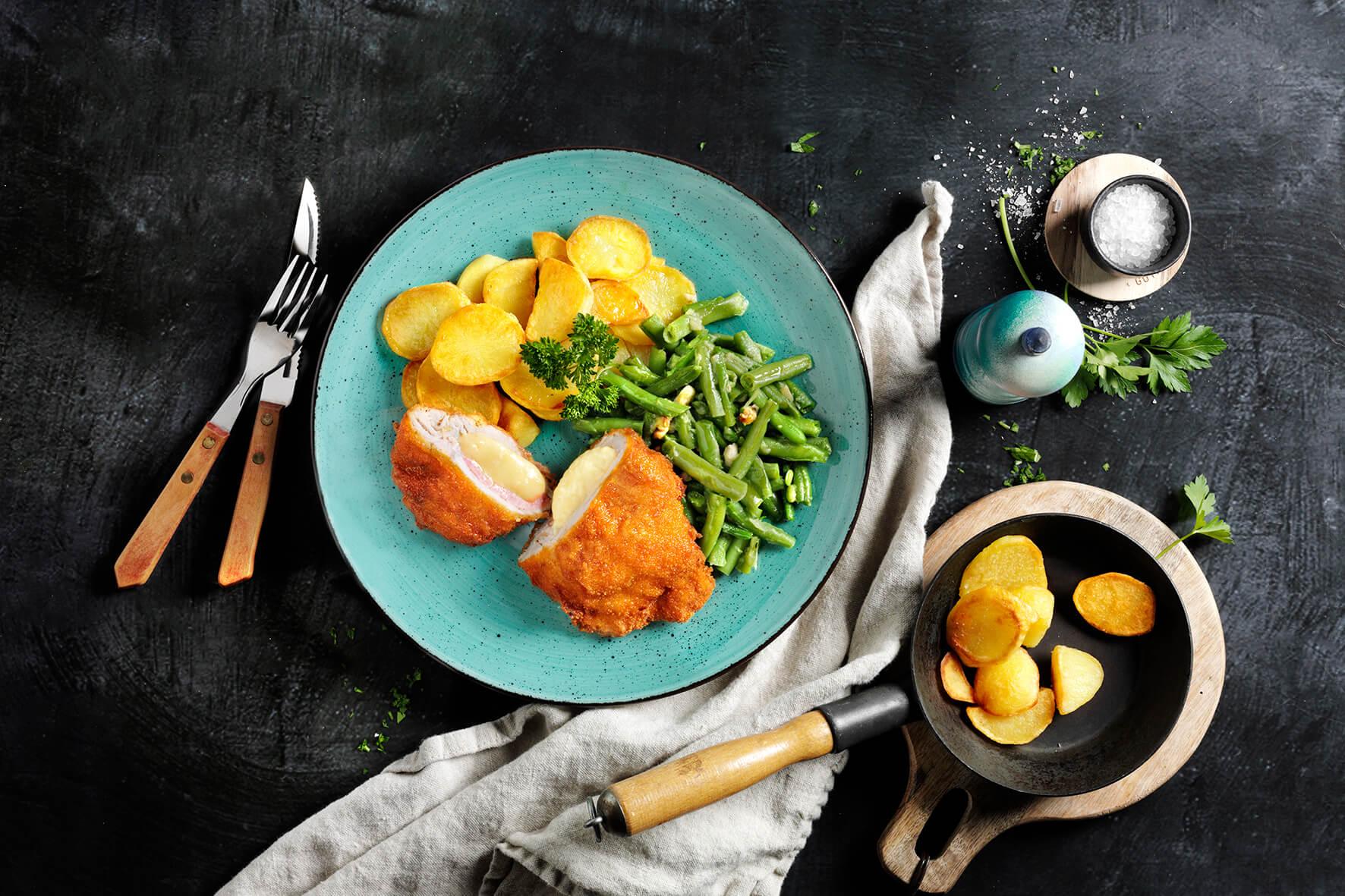 HOFMANNs Schnitzel-Box: paniertes Hähnchen, grüne Bohnen und Bratkartoffeln, Besteck liegt bereit.
